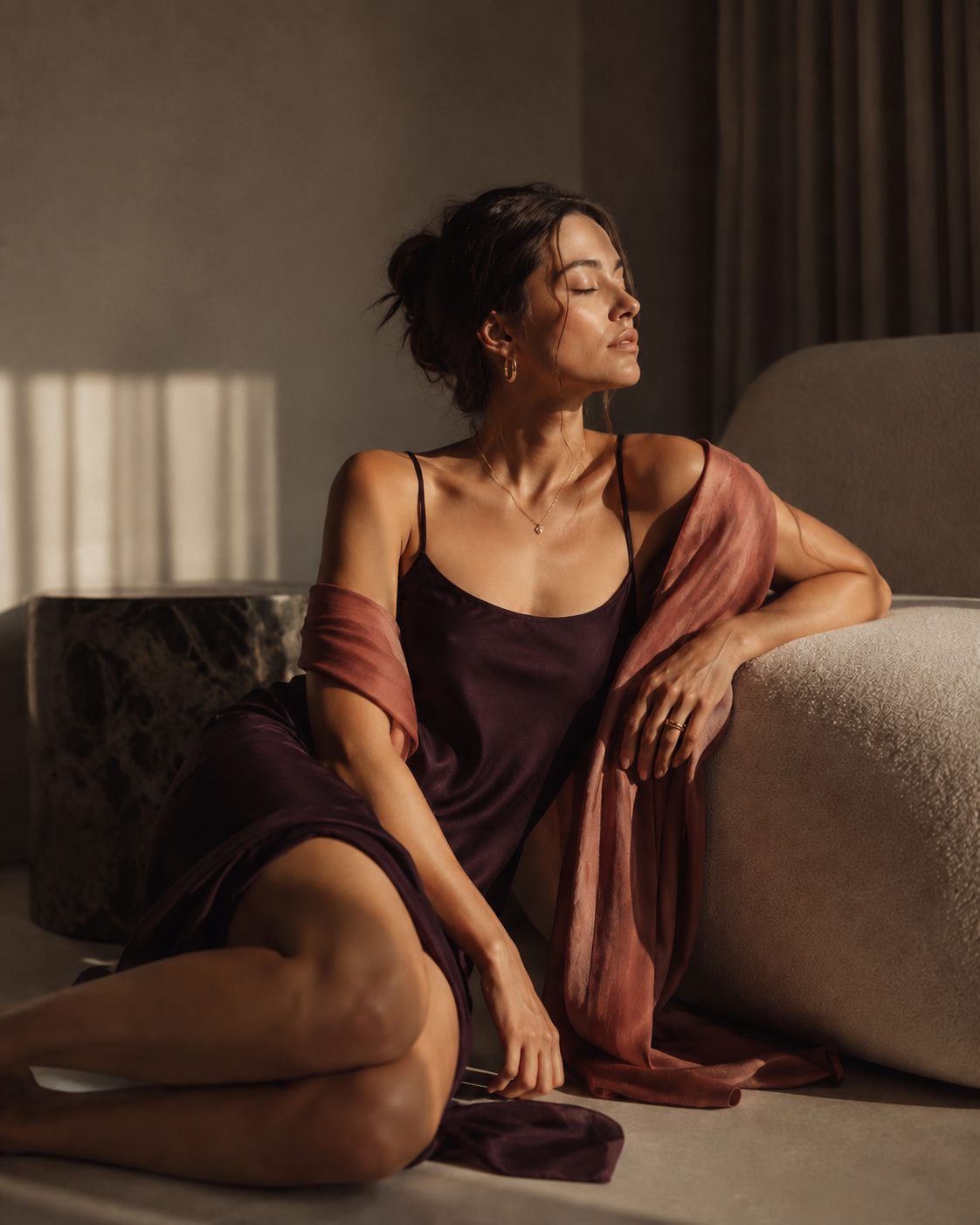 Woman in a plum silk slip and wrap relaxing against a cream sofa in warm evening light